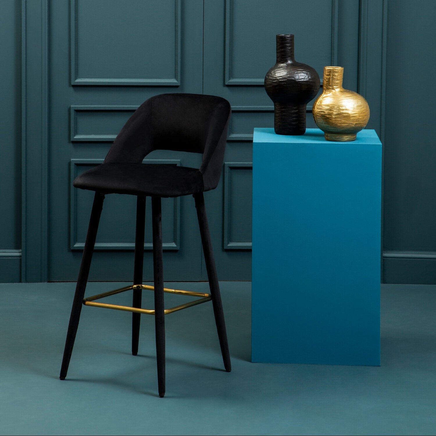 Black bar stool in front of a blue pedestal with two vases against a teal wall.