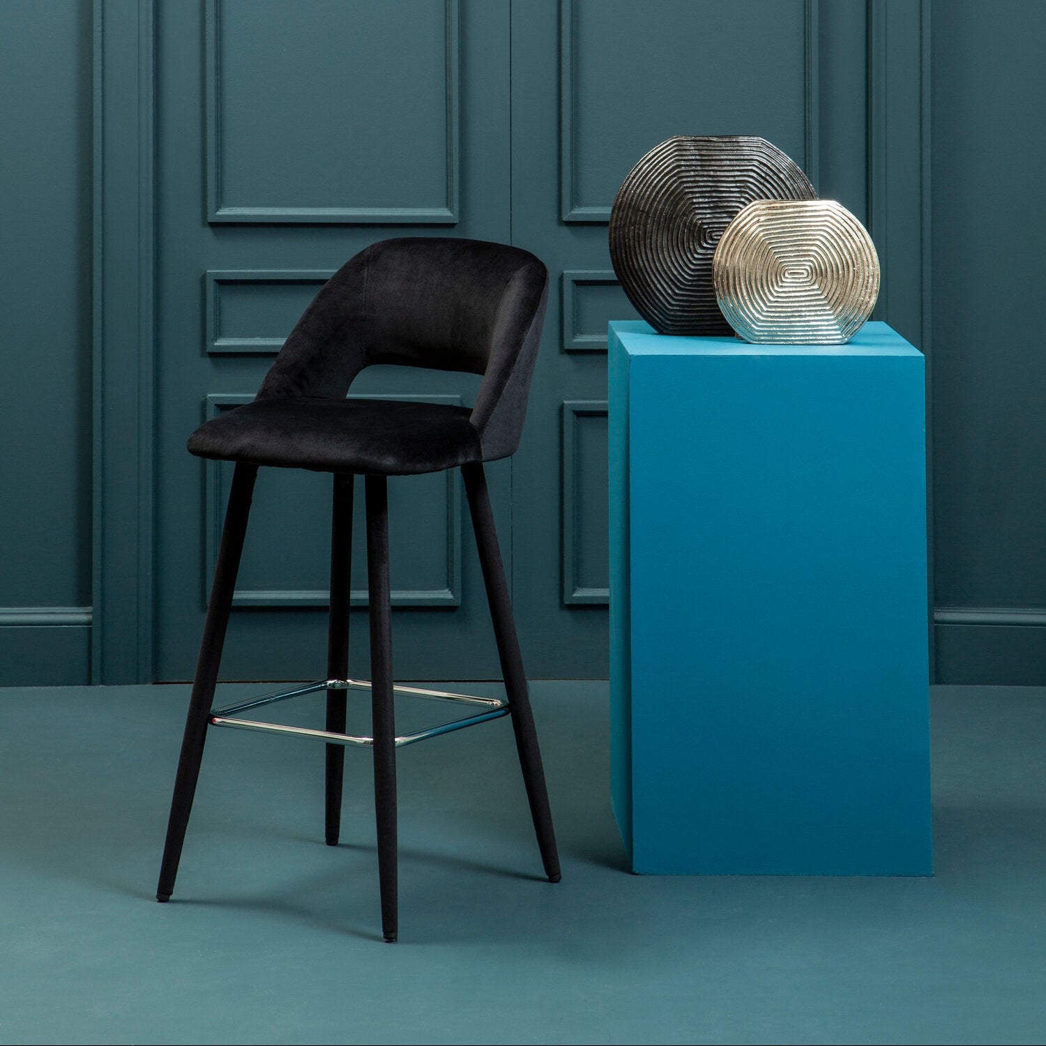 Dark brown bar stool in front of a teal wall with a blue pedestal holding decorative items.