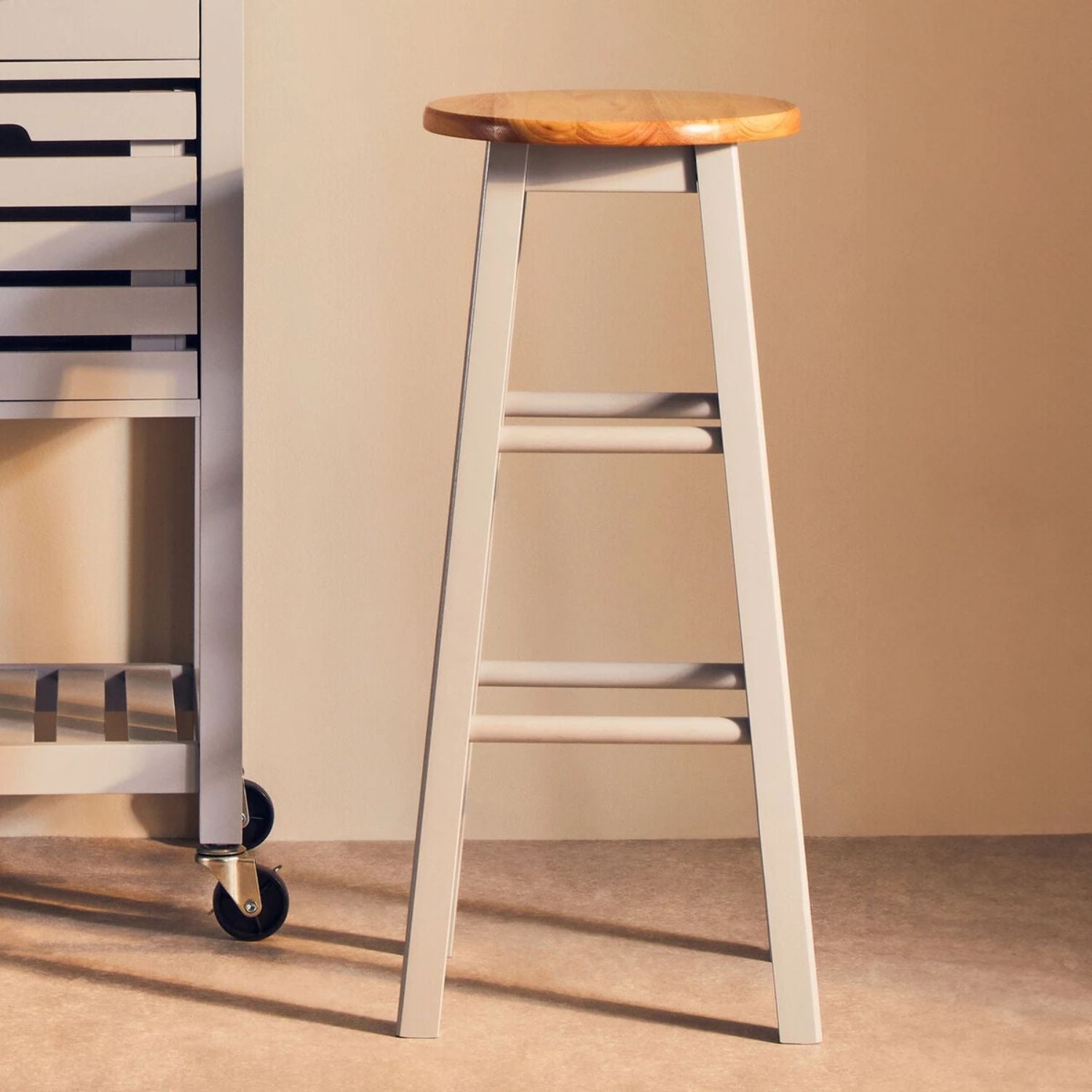 Bar stool with wooden seat and white legs against a beige wall.