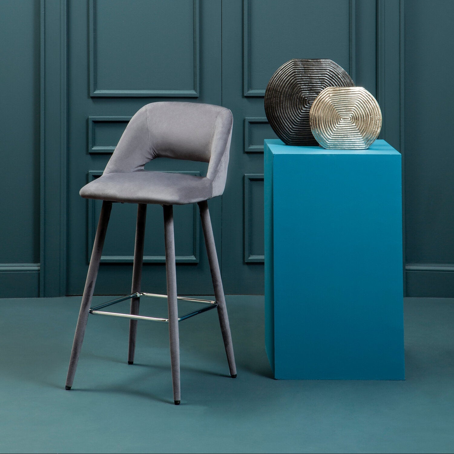 Gray bar stool next to a blue pedestal with decorative items against a teal wall.