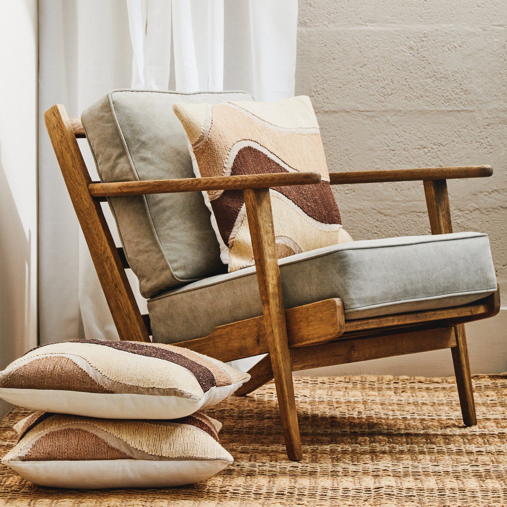 Wooden armchair with patterned cushions in a room with light curtains and a textured wall.