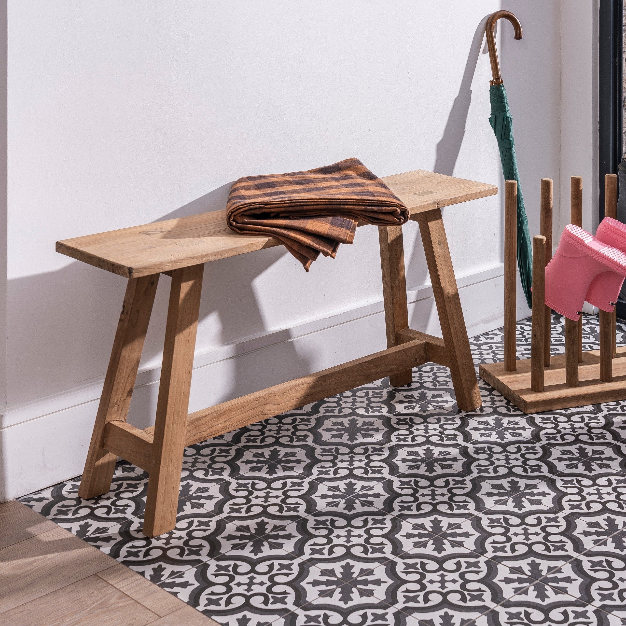 Wooden bench with folded coat, umbrella stand with pink shoes, and boots on a patterned floor.
