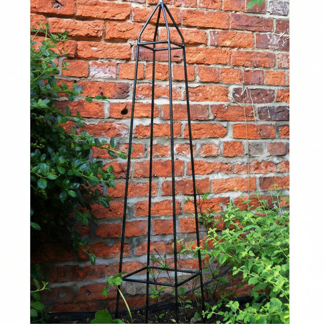 Black metal garden obelisk against a brick wall with greenery